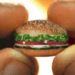 Tiny hamburger being held by giant fingers.