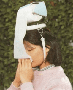 Photo of person with a toilet paper holder for a hat and blowing their nose with some of the toilet paper. 