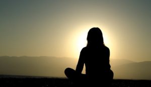 Silhouette of a person sitting on a mountain in a meditation pose as the sunsets behind a mountain top. 